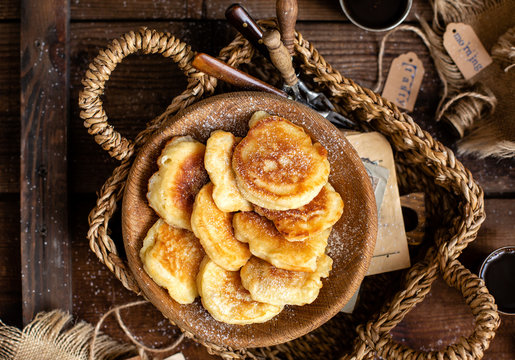 overhead breakfast shot with tasty fried russian pancakes with powdered sugar on top in wooden bowl stands in wicker straw basket with forks on rustic brown table