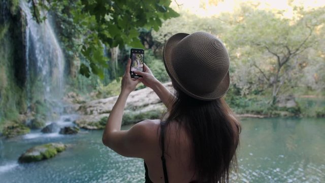 Rear view of woman in hat taking photograph smartphone sharing photo of waterfall nature background.