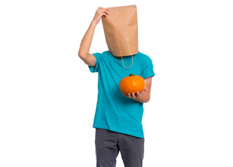Happy halloween concept. Teen boy with paper bag over his head holds orange small fresh pumpkin, isolated on white background. Teenager cover head with bag. Child with food - raw vegetables in hands.