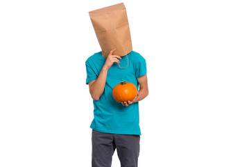 Happy halloween concept. Teen boy with paper bag over his head holds orange small fresh pumpkin, isolated on white background. Teenager cover head with bag. Child with food - raw vegetables in hands.