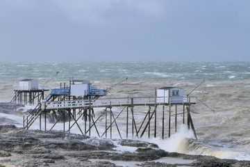Fototapeta na wymiar Pier in the sea. White fisherman houses stand on the coast. Storm, stormy sea and high waves can be seen. It is an uncomfortable day in the afternoon. No people are visible.