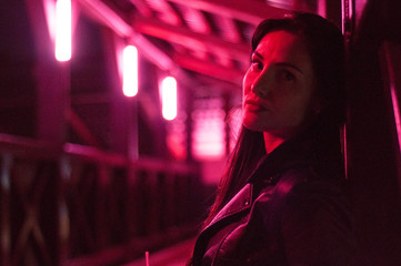 A young girl with long black hair in a black jacket posing in a long corridor with pink light