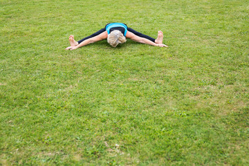 Fototapeta na wymiar A middle aged woman practicing yoga barefoot outside in a grassy park. She is wearing a bright blue vest and black leggings. The style of yoga she is doing is Hatha Yoga