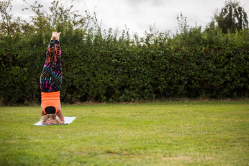 A middle aged woman practicing yoga barefoot outside in a grassy park. She is wearing a bright orange vest and multi coloured leggings. The style of yoga she is doing is Hatha Yoga