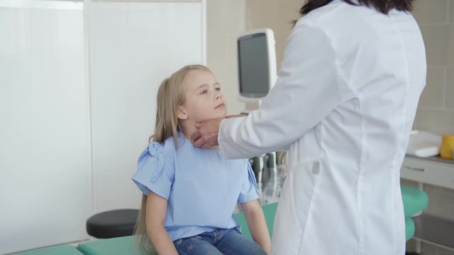 Medium tracking shot of female pediatrician in white coat checking tonsils and throat of smiling little girl sitting on couch in doctors office