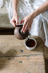 Fototapeta na wymiar Selective focus of young adult woman holding teapot and pouring tea in white cup. Girl resting on weekend holiday, sitting near wooden table on sofa with plaid and spending free time at cozy home