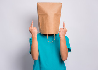 Portrait of teen boy with paper bag over head making middle fingers. Teenager cover head with bag showing bad gesture posing in studio. Child pulling paper bag over head doing obscene sign.