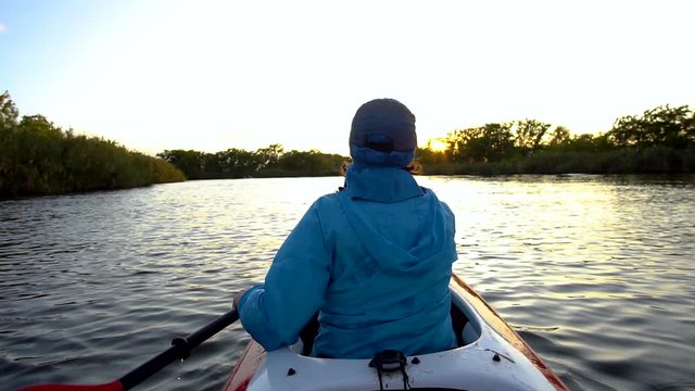 A woman is swimming in a kayak towards the setting sun on a calm river. She is rowing with oars. Slow motion