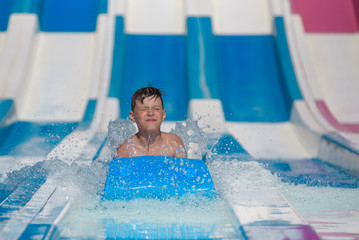 Cute boy sliding on floater down slide in waterpark. He enjoying his summer vacations. - 291564341