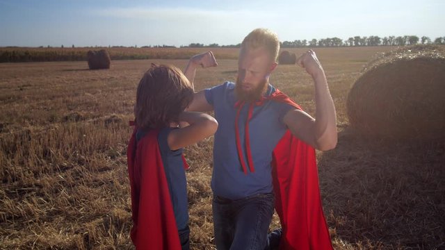 Athletic man with red hair and beard teaching son to show biceps muscles while playing superheroes on wheat field at sunset. Cute little boy feeling impressive father's biceps wanting to be as strong