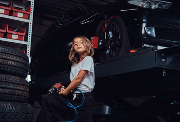 Little cute girl is posing for photographer in dark auto workshop with pneumatic drill in hands.