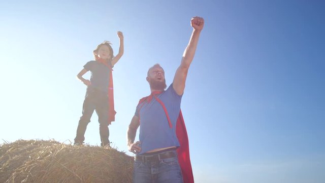 Serious dad and son playing superheroes while stretching clenched fist forward and up. Little boy standing on straw stack near father during play on background of blue sky