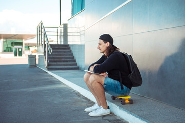 A young man in a baseball cap, with a backpack, and in denim shorts, drinks coffee while sitting on a yellow skateboard.