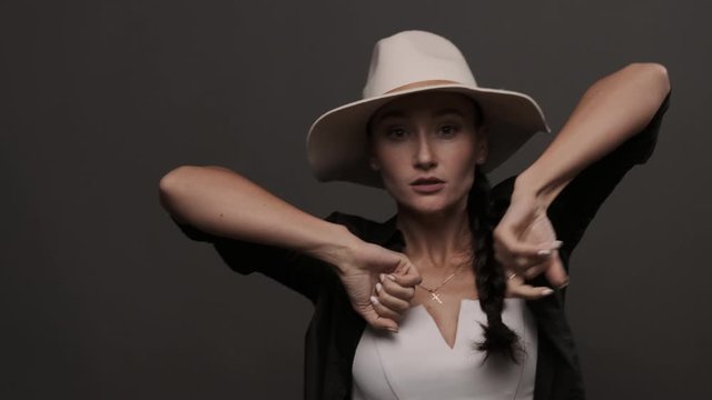 Stylish girl in hat performing cool dance and looking at camera. Caucasian woman dancing in studio. Contemporary dance movement. Listening to music. Female dancer enjoying modern waacking dance style
