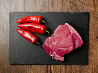 Portion of raw Sirloin steak plate and three red chilly peppers on a slate on a wooden table, Top view.