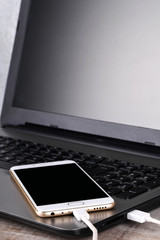 A gray laptop and a white telephone(smartphone) connected by a cable for data transmission on a wooden table. Data transfer. Information exchange