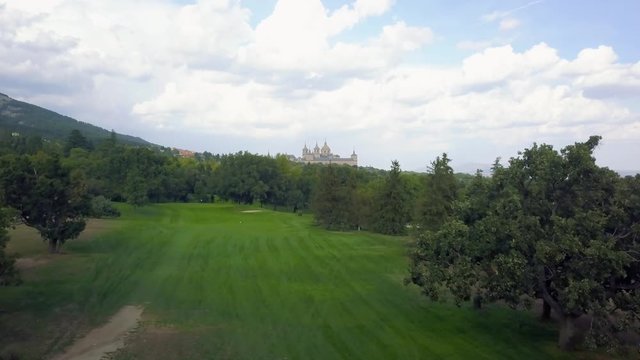 golf course in a forest of ash with the historic monastery of escorial crowning a hill, aerial view over hills