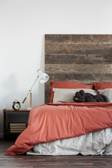 Trendy white lamp next to black clock on wooden nightstand next to king size bed with wooden headboard