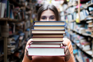 girl student holds a stack of books in the library, she searches for literature and offers to read, a woman prepares for study, knowledge is power, bookseller on the background of the bookstore