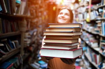 girl student holds a stack of books in the library, she searches for literature and offers to read, a woman prepares for study, knowledge is power, bookseller on the background of the bookstore