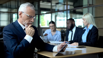 Mature businessman thinking of decision looking tablet, managers discussing plan