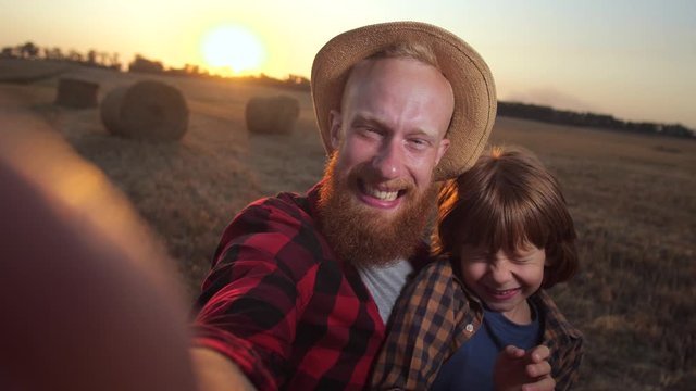 Close-up portrait of redhead father and son making funny faces while taking photo on smartphone against background of countryside landscape. Happy family posing for selfie in the middle of wheat field