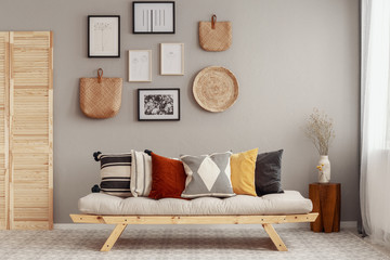 Dry flowers in white vase on small wooden table behind comfortable futon sofa with pillows