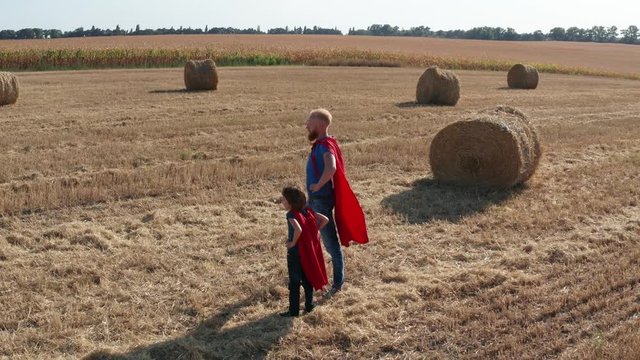 Aerial view of superheroe family looking out for danger and rushing to rescue while holding hands and stretching fist forward. Drone shot of father and son dressed as supermen running across field