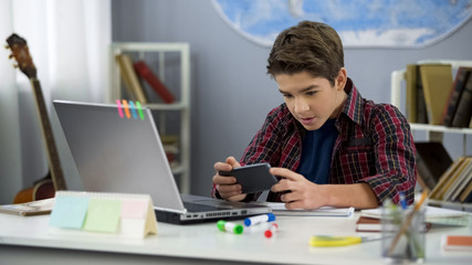 Male school pupil watching online video holding smartphone sitting home desk - obrazy, fototapety, plakaty