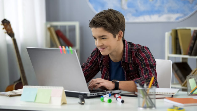 Smiling teen male chatting with friends in social networks sitting front laptop