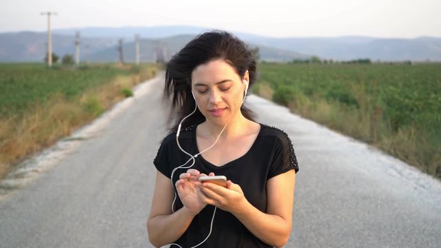 Relaxed young woman wearing a black t-shirt listening to the music and walking along a river bank. Tracking real time establishing shot
