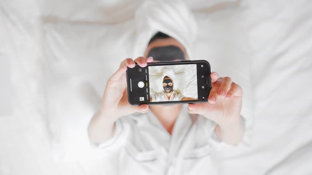 woman with a white towel on her head and black cosmetic mask lying on the bed using smartphone take photo at home wellness, spa, skin care