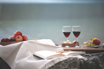 2 glasses of red wine, an open book, a basket with fruits and salami and cheese on a stone with yellow leaves on a lake background