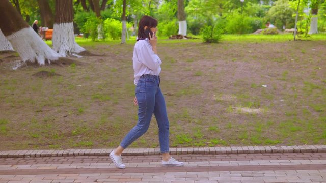 side view young woman in white shirt and denim talking by phone walking on the street in summer season businesswoman walking with device on the background green nature in the park
