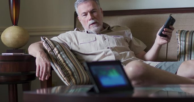 A mature man relaxing on a couch looking at his smartphone asks his smart home voice controlled device something and reacts to the response.