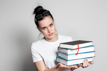 Young, puzzled woman student with glasses on head holds a stack of books in her hands and looks thoughtfully at the camera, on a white background.