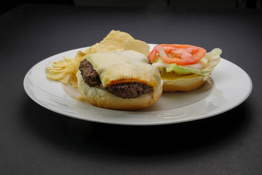 Homestyle open-face cheeseburger on bun with lettuce tomato, onion, and potato chips served on white plate with dark background.