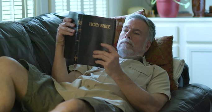 A Middle Aged Caucasian Man Relaxing On His Couch Reading From A Large Traditional Style Christian Bible Gets Sleepy And Falls Asleep.