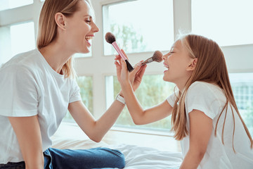 Spending time together. Happy delighted mother and her little adorable daughter are standing and holding brushes while doing make-up for each other