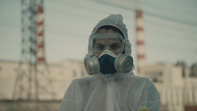 The scientist put on a respirator and a protective gown. Girl student and a stern look on the background of the plant. Atmospheric pollution. Coronavirus in a pandemic, COVID-19.