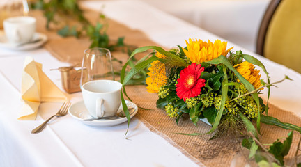 Festive table setting. Coffee cups, glasses, bouquets of sunflowers and red gerberas, on a white tablecloth.
