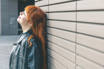 Fototapeta premium Cute young girl in a black leather jacket with curly red hair and a fringe relaxes near a modern wall and enjoys a sunny day.