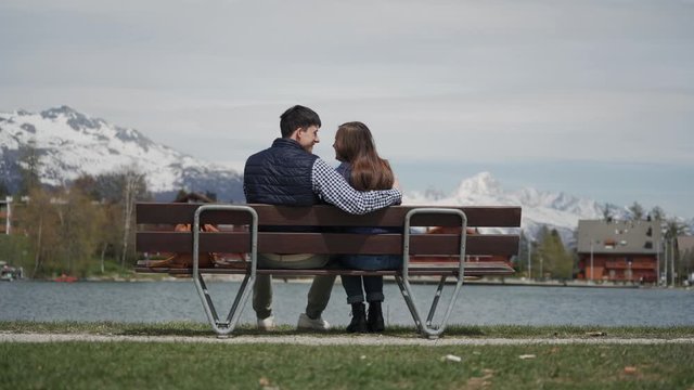 Happy young couple sitting on lakeside wooden bench with beautiful view on Swiss Alps. Guy hugs his girlfriend while relaxing at nature. Rear view
