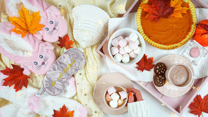 Cozy autumn Thanksgiving in bed flatlay overhead with cosy sweater, slippers, socks and tray with pumpkin pie and hot chocolate with marshmallows.