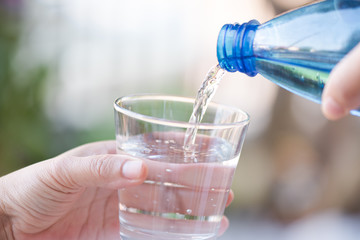 Hand holding blue water bottle pouring water into the glass on blurry natural green tree background.Health care food and drink concept.