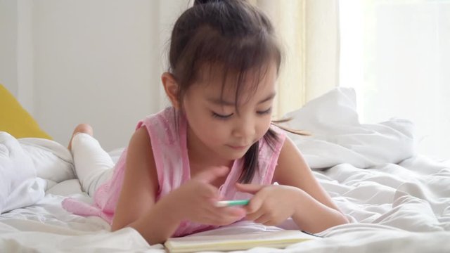 4K Medium shot happy smiling little cute asian preschool child girl lying down on the bed enjoy doing homework or writing on the book. Relaxing learning homeschool education and back to school concept