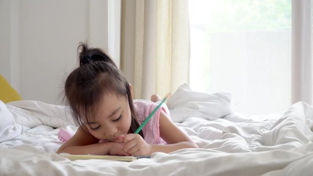 4K Medium shot happy smiling little cute asian preschool child girl lying down on the bed enjoy doing homework or writing on the book. Relaxing learning homeschool education and back to school concept