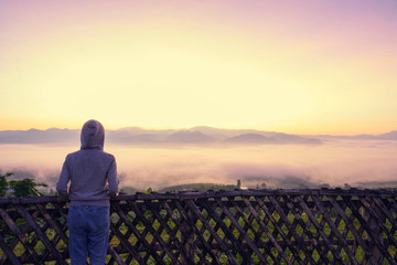Woman with hood jacket cover head standing behind the fench on the top of the mountain looking out misty in the valley early morning twilight sky before sunrise sky