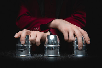 Magician shows shell game of thimbles with circles and ball, black background. Concept deception,...