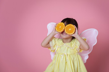 Little girl in angel dress with white wing playing with fresh orange fruits on pink background,Healthy eating concept.